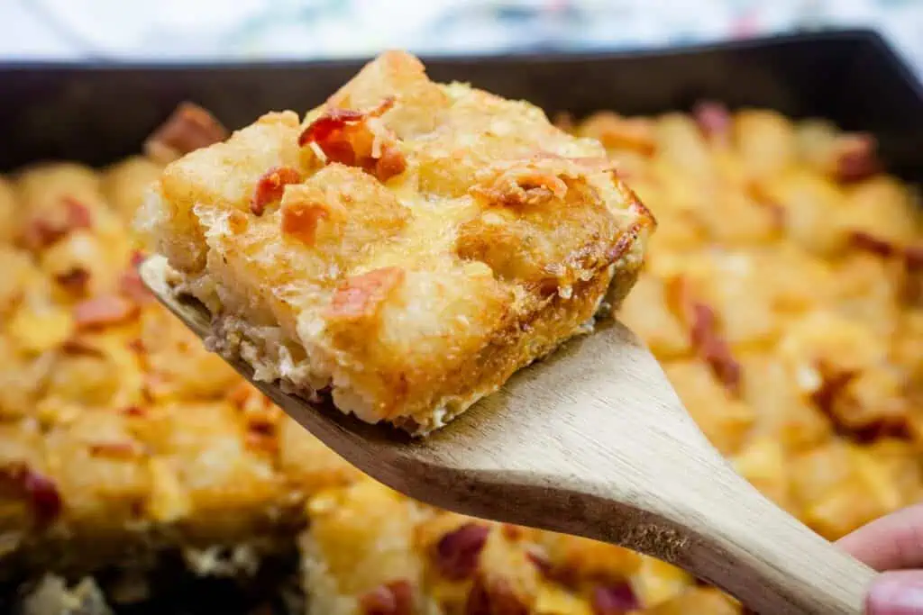 A wooden spatula holds a square piece of baked casserole topped with melted cheese and bits of bacon, with more casserole visible in the background.