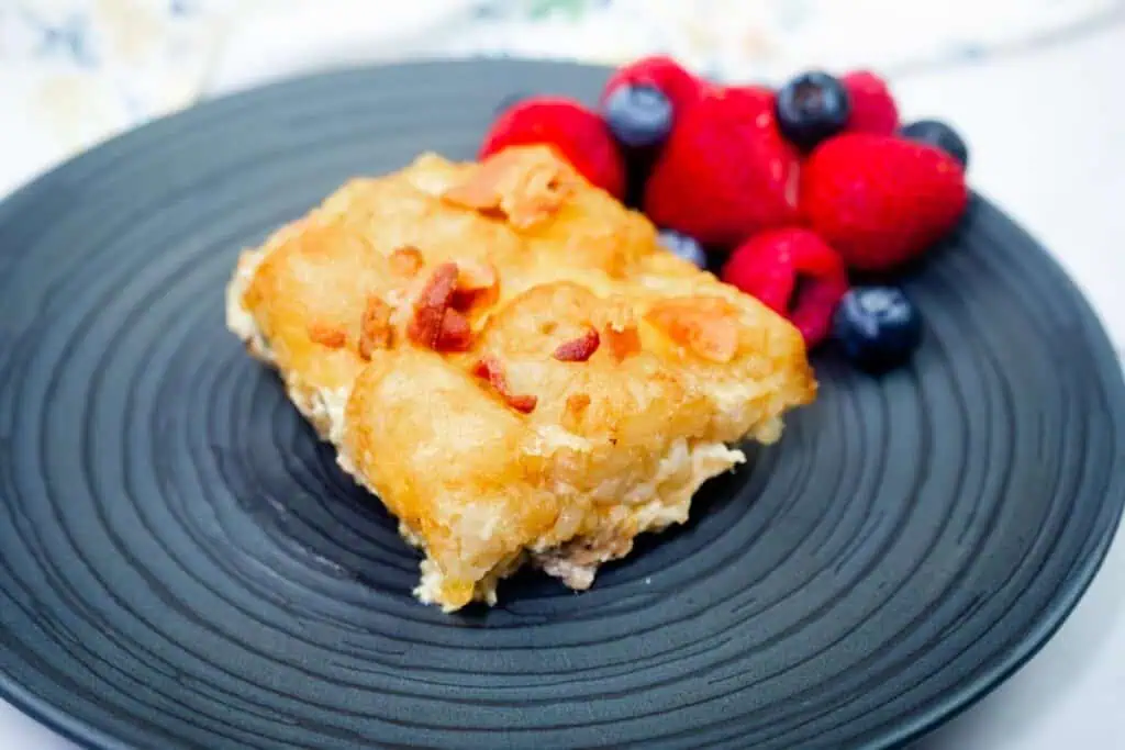 A serving of breakfast casserole topped with bacon sits on a black plate next to fresh raspberries, strawberries, and blueberries.
