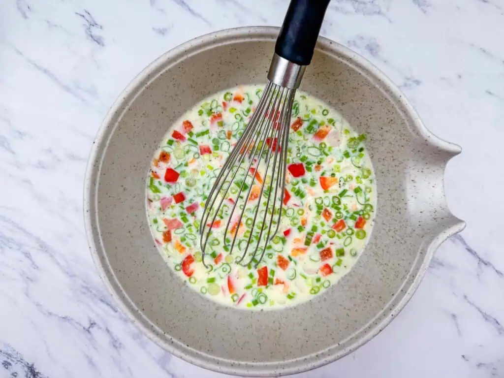 A mixing bowl with chopped vegetables, eggs, and milk being whisked together, sitting on a marble surface.