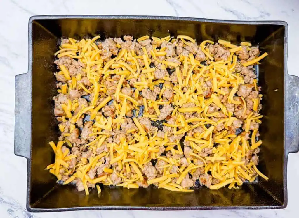 Rectangular baking dish with a layer of cooked ground meat and shredded cheddar cheese on top, placed on a white marble surface.