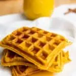 A stack of three square waffles with syrup on a white plate, with a glass of orange juice in the background.