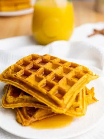 A stack of three square waffles with syrup on a white plate, with a glass of orange juice in the background.