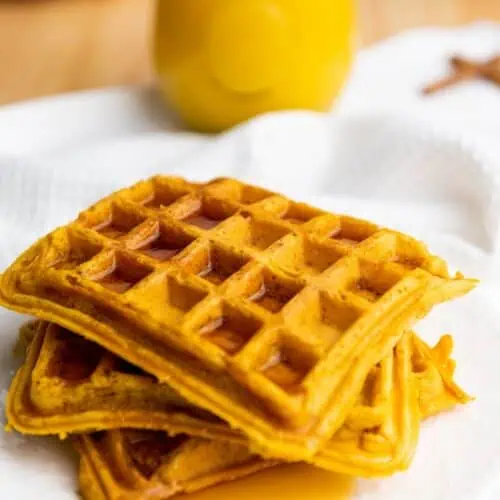 A stack of three square waffles with syrup on a white plate, with a glass of orange juice in the background.