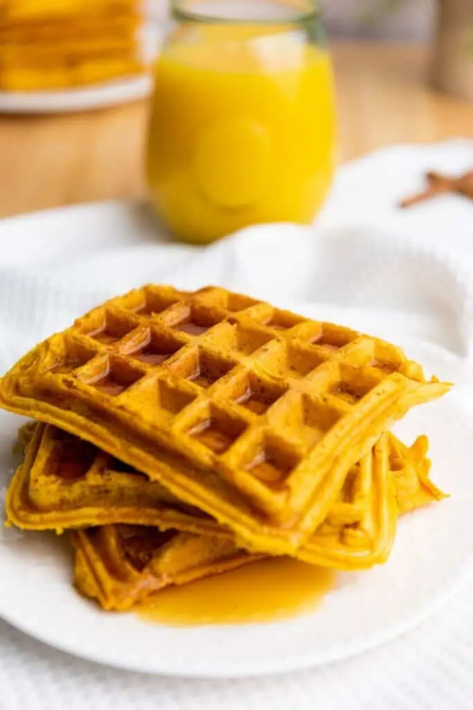 A stack of three square waffles with syrup on a white plate, with a glass of orange juice in the background.