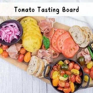 A wooden board with sliced tomatoes, cherry tomatoes, prosciutto, sliced bread, pickled red onions, and basil, labeled "Tomato Tasting Board.