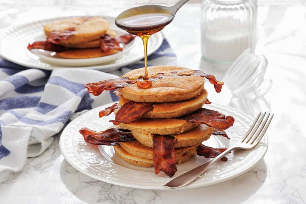 A stack of Bacon Pancakes on a white plate, with syrup being poured on top and a fork beside the plate. A jar of milk and another plate are in the background.