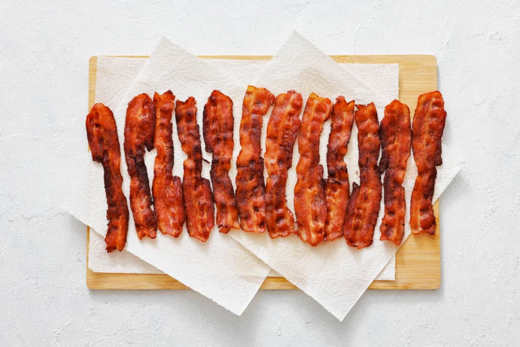 Crispy strips of cooked bacon are arranged in a row on paper towels placed on a wooden cutting board.