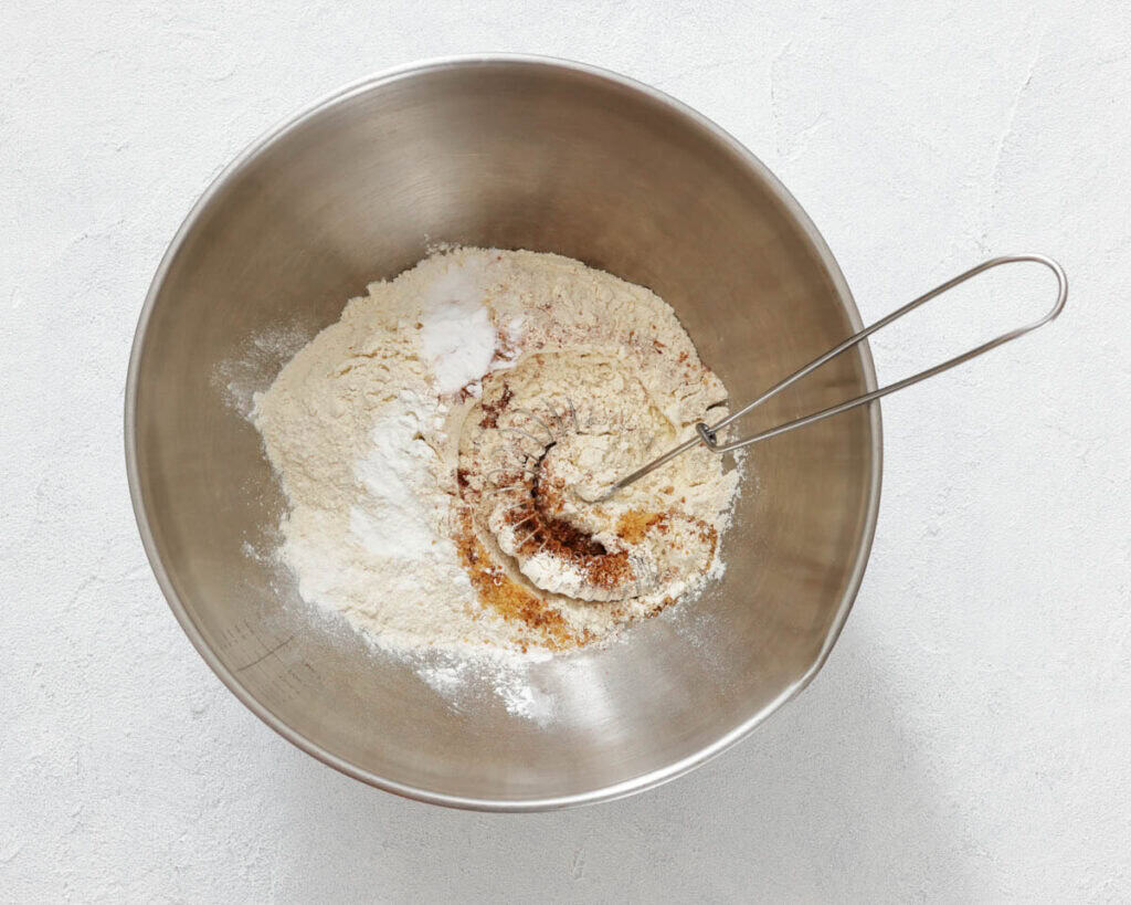 A metal mixing bowl contains flour, baking soda, baking powder, cinnamon, and other dry ingredients, with a metal whisk resting inside.