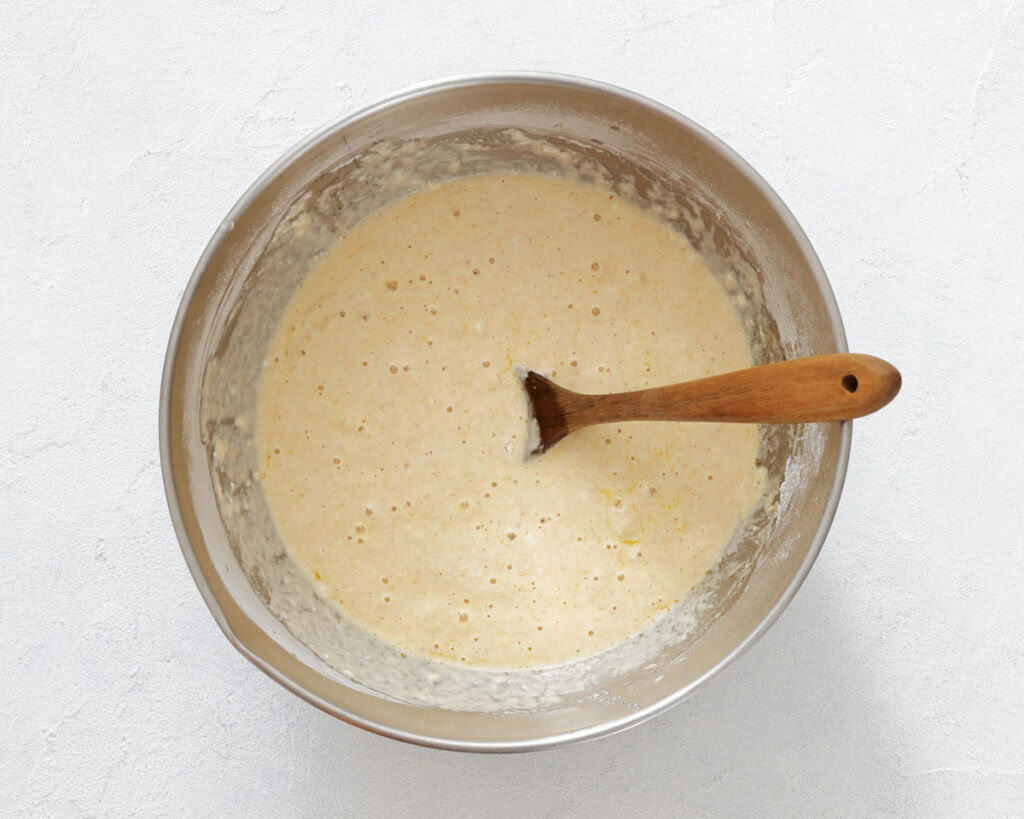 A stainless steel bowl filled with pancake batter sits on a white surface with a wooden spoon partially submerged in the mixture.