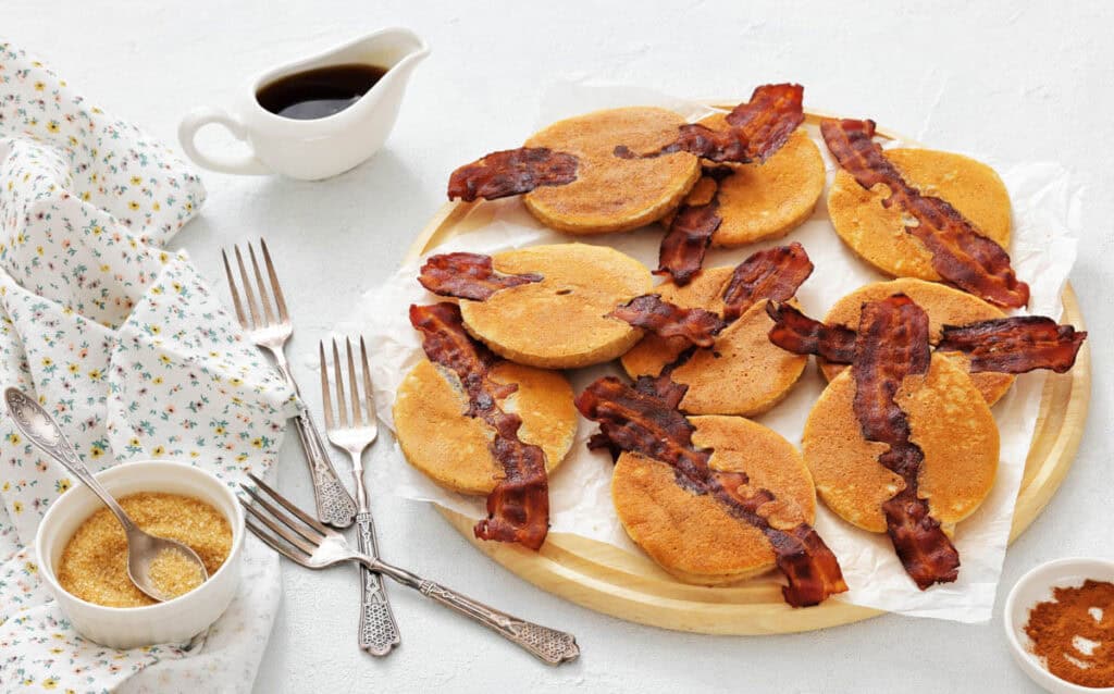 A round wooden tray holds nine Bacon Pancakes on a parchment paper. A small pitcher of syrup, forks, and bowls of brown sugar and cinnamon are nearby.