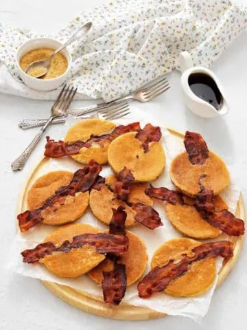 A wooden tray with mini pancakes each topped with a strip of crispy bacon, with syrup and brown sugar in small containers, and a floral napkin and forks nearby.