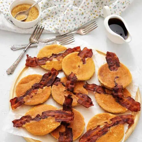 A wooden tray with mini pancakes each topped with a strip of crispy bacon, with syrup and brown sugar in small containers, and a floral napkin and forks nearby.