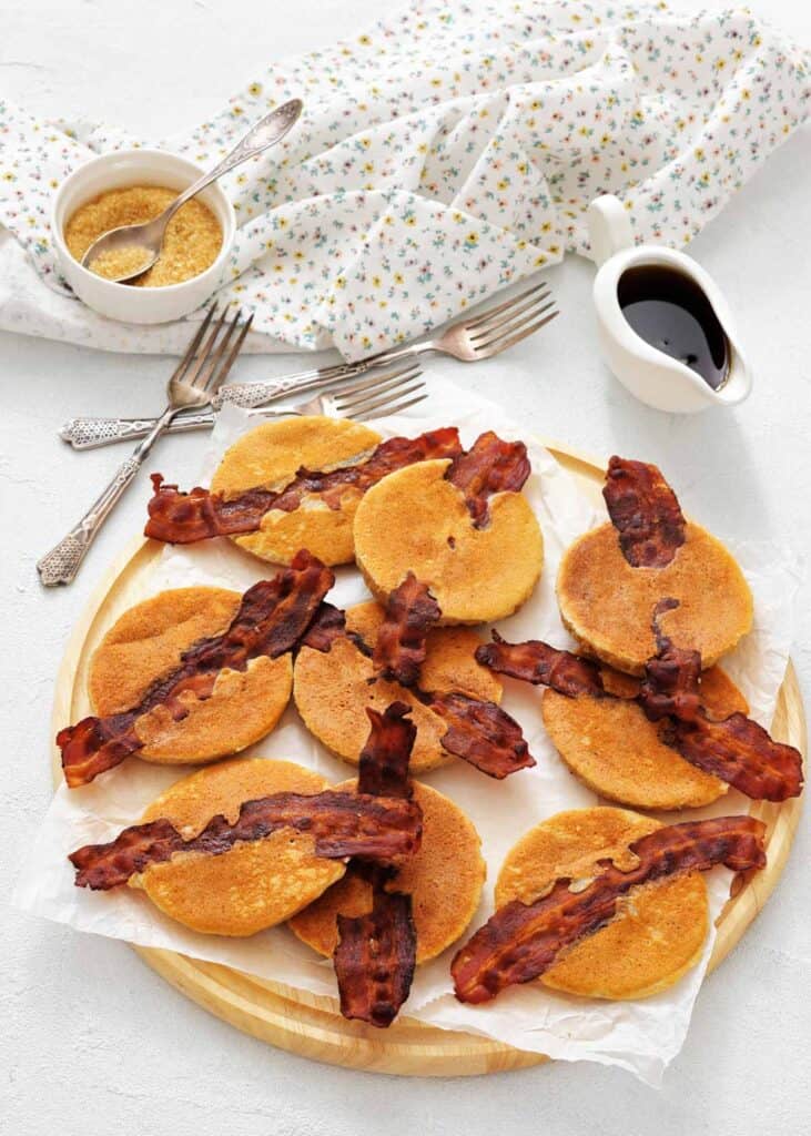 A wooden tray with mini pancakes each topped with a strip of crispy bacon, with syrup and brown sugar in small containers, and a floral napkin and forks nearby.