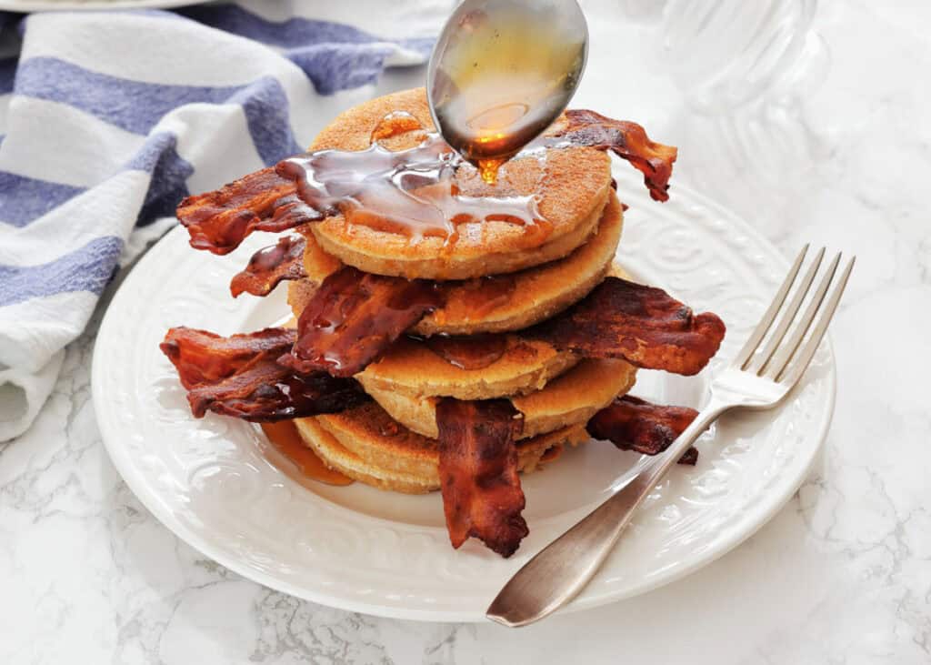 Stacked on Bacon Pancakes on a white plate, being drizzled with syrup from a spoon, with a fork beside the stack.