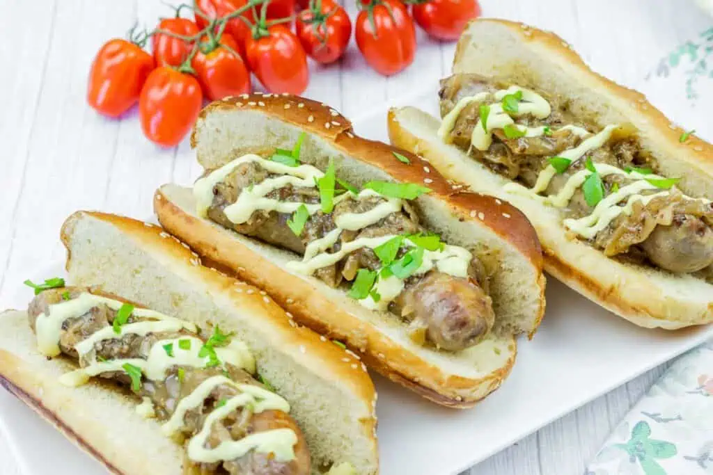 Three sausage sandwiches in buns topped with chopped herbs and sauce, served on a white plate with cherry tomatoes in the background.