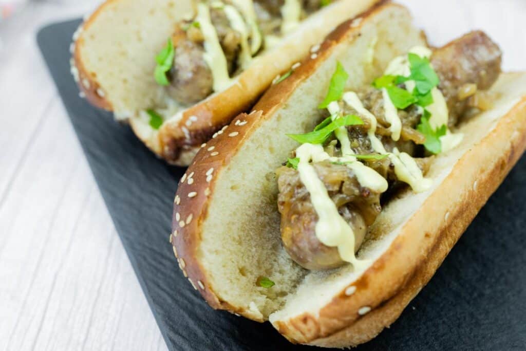 Two sausage sandwiches in sesame seed buns, topped with saut&eacute;ed onions, drizzled with creamy sauce, and garnished with fresh parsley on a black tray.