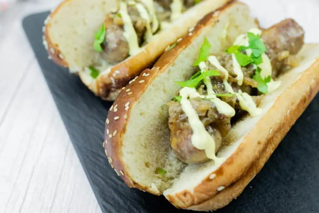 Two sausage sandwiches in sesame seed buns, topped with sautéed onions, drizzled with creamy sauce, and garnished with fresh parsley on a black tray.