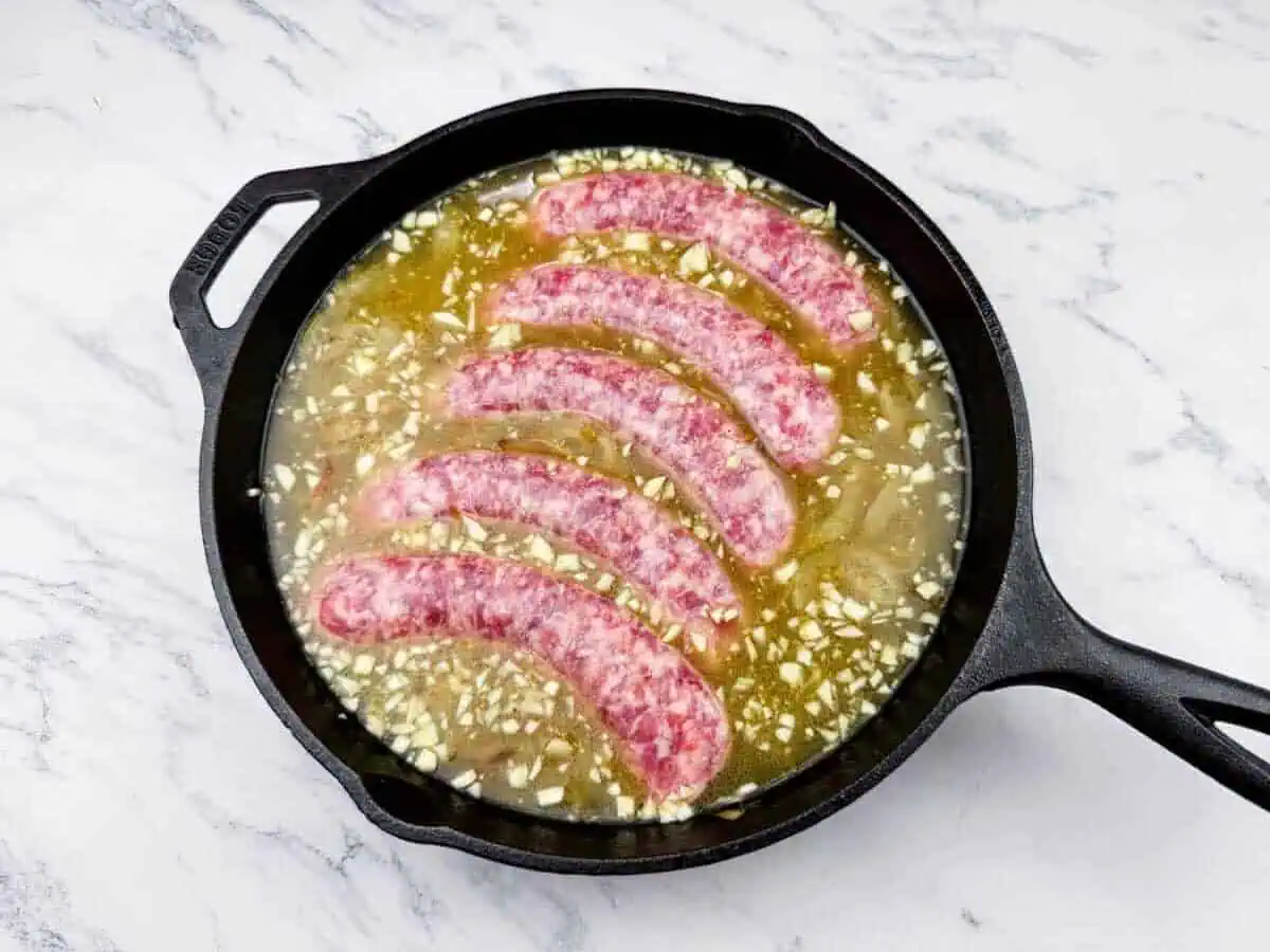 Five raw sausages in a cast iron skillet with chopped garlic and liquid, placed on a white marble surface.