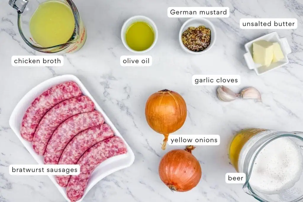 Ingredients for a bratwurst dish arranged on a marble surface: bratwurst sausages, chicken broth, olive oil, German mustard, unsalted butter, garlic cloves, yellow onions, and beer.