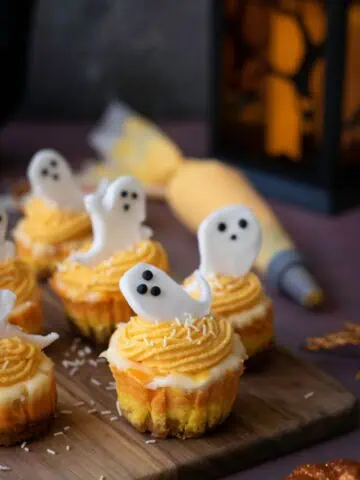 Mini cupcakes with orange frosting and white ghost-shaped decorations are arranged on a wooden board, with a piping bag and lantern in the background.