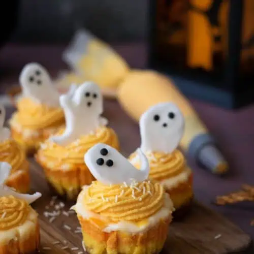 Mini cupcakes with orange frosting and white ghost-shaped decorations are arranged on a wooden board, with a piping bag and lantern in the background.