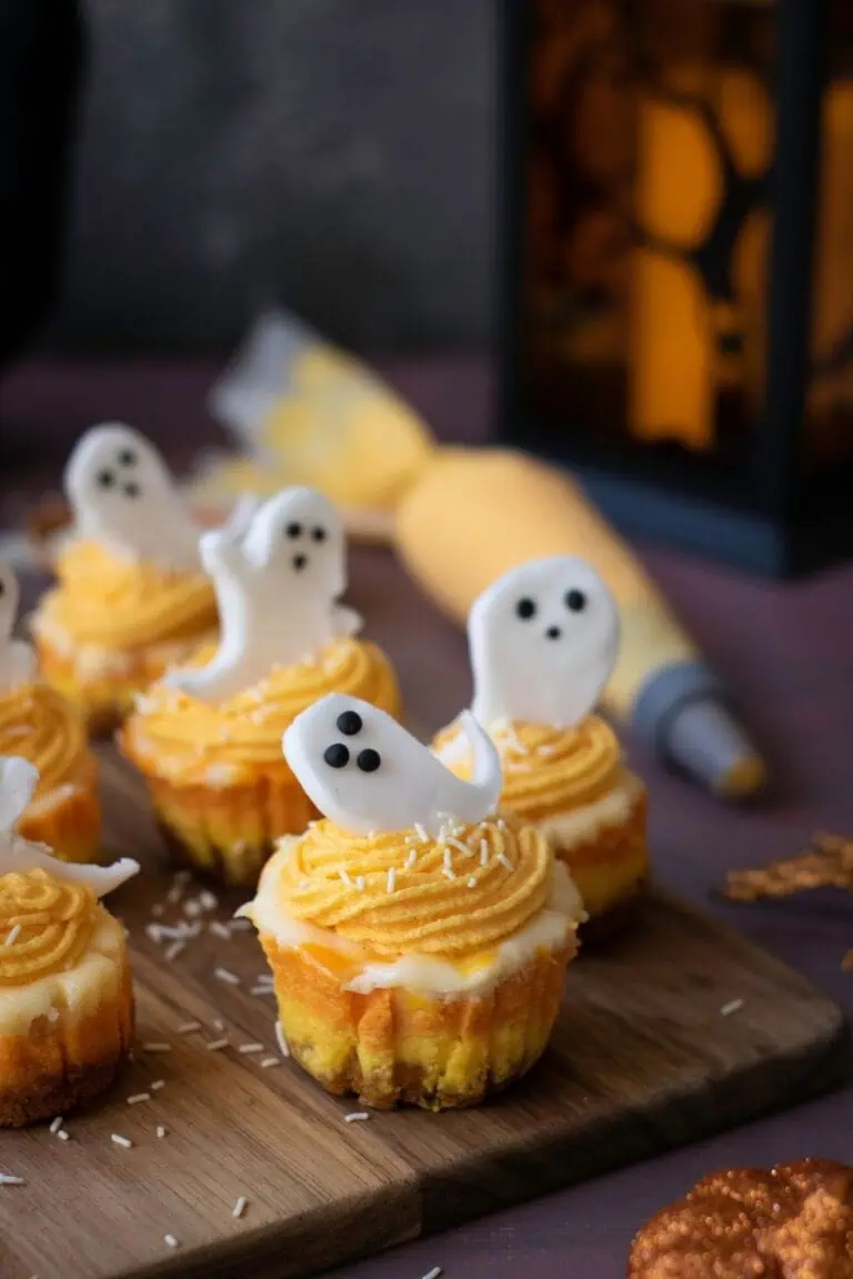 Mini cupcakes with orange frosting and white ghost-shaped decorations are arranged on a wooden board, with a piping bag and lantern in the background.