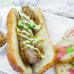A sausage in a bun topped with sautéed onions and a drizzle of sauce, served on a white plate with sliced potatoes and herbs.