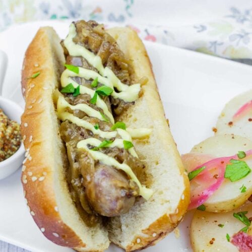 A sausage in a bun topped with saut&eacute;ed onions and a drizzle of sauce, served on a white plate with sliced potatoes and herbs.