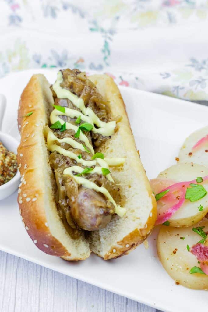 A sausage in a bun topped with saut&eacute;ed onions and a drizzle of sauce, served on a white plate with sliced potatoes and herbs.