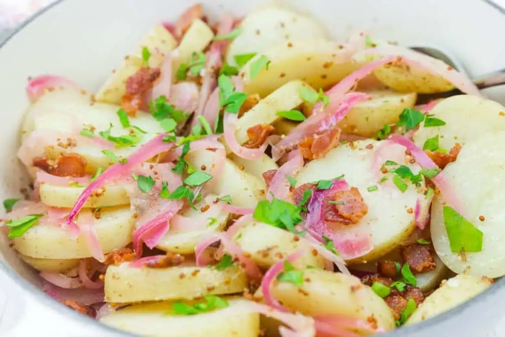 A bowl of sliced potatoes mixed with bacon, red onions, chopped parsley, and a grainy mustard dressing.