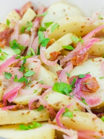 A bowl of sliced potatoes mixed with bacon, red onions, chopped parsley, and a grainy mustard dressing.