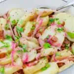A white dish filled with sliced potatoes, red onions, bacon bits, and chopped parsley, with a serving spoon on the side.