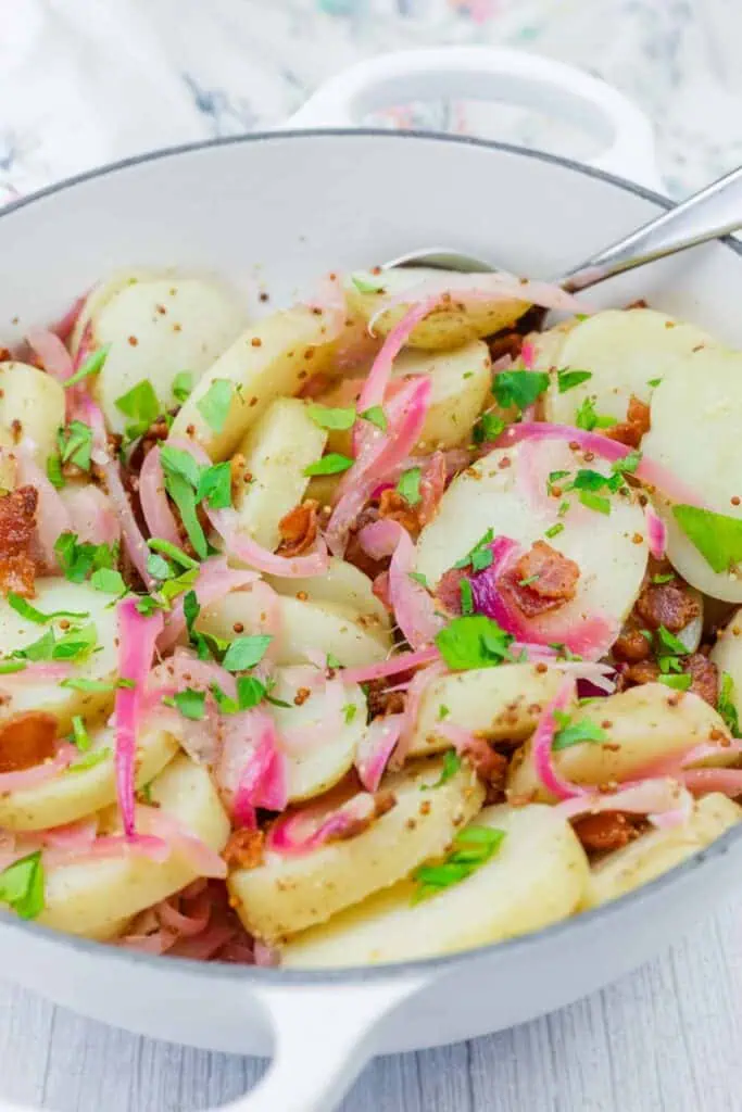 A white dish filled with sliced potatoes, red onions, bacon bits, and chopped parsley, with a serving spoon on the side.