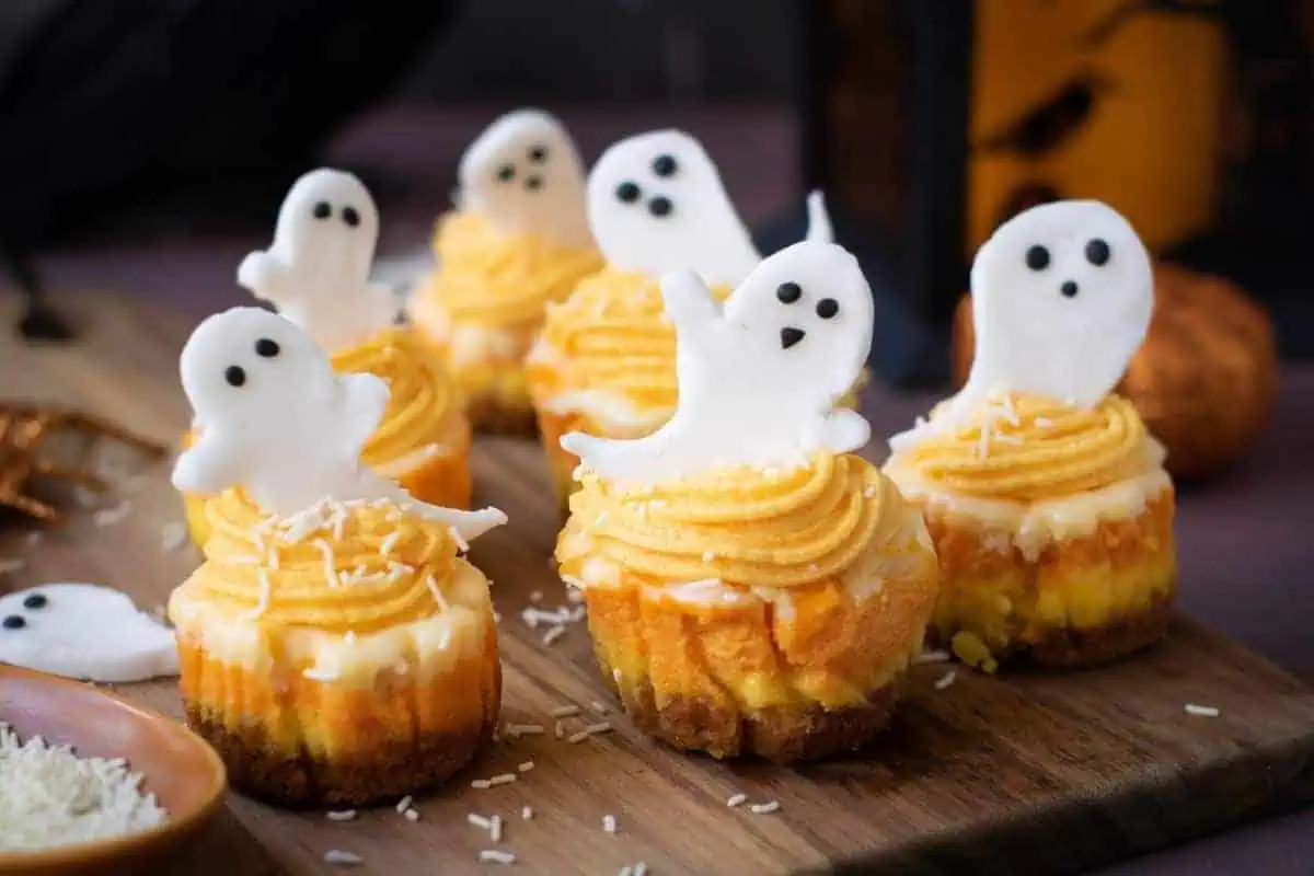 Mini cheesecakes topped with yellow frosting and white fondant ghost decorations are arranged on a wooden board for a Halloween-themed display.