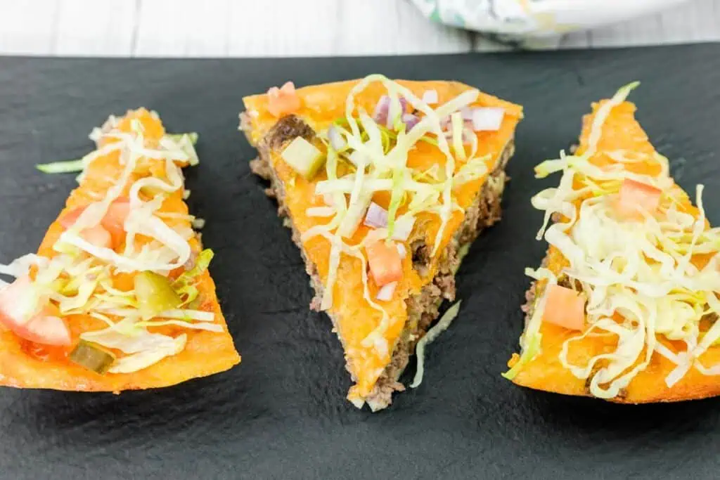 Three slices of cheeseburger pie topped with ground beef, shredded lettuce, diced tomato, onion, and pickles are arranged on a black plate.