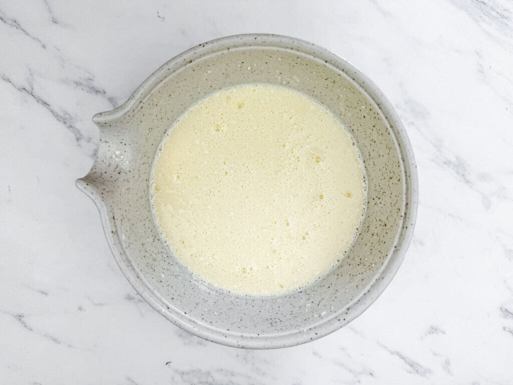 A speckled grey mixing bowl containing the mixture of milk, Bisquick, eggs, salt, pepper, and garlic powder sits on a white marble surface.