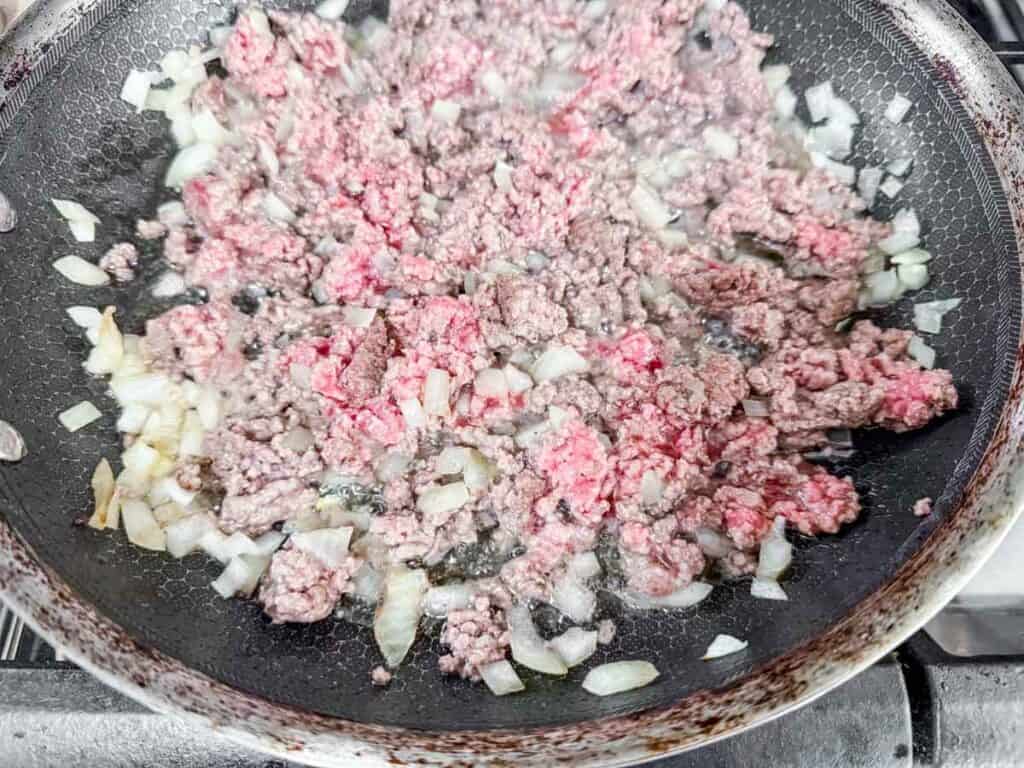 Ground beef and chopped onions cooking in a black frying pan on a stove.