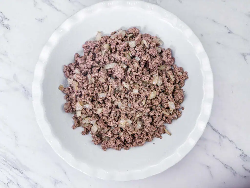 A white plate containing cooked ground beef mixed with diced onions, placed on a marble surface.