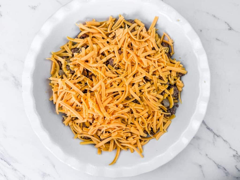 A white pie dish filled with shredded cheddar cheese and cooked ground beef, on a marble surface.