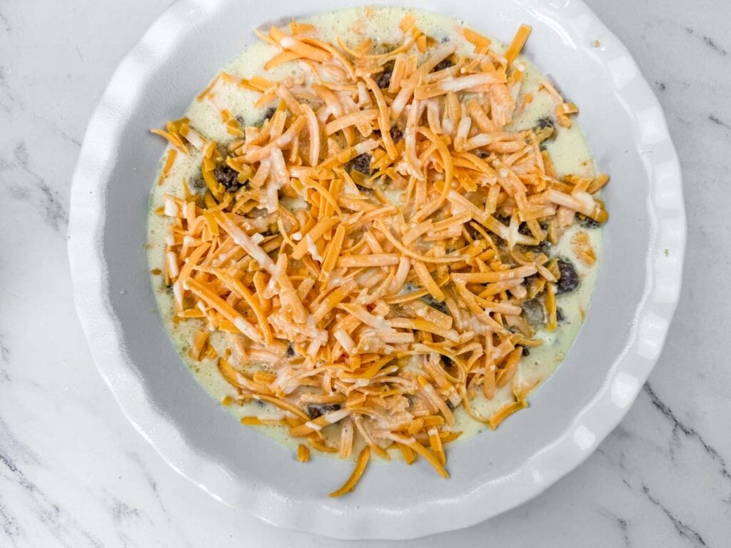 A pie dish filled with a mixture of cooked ground beef, creamy batter, and shredded cheddar cheese on a white marble surface.
