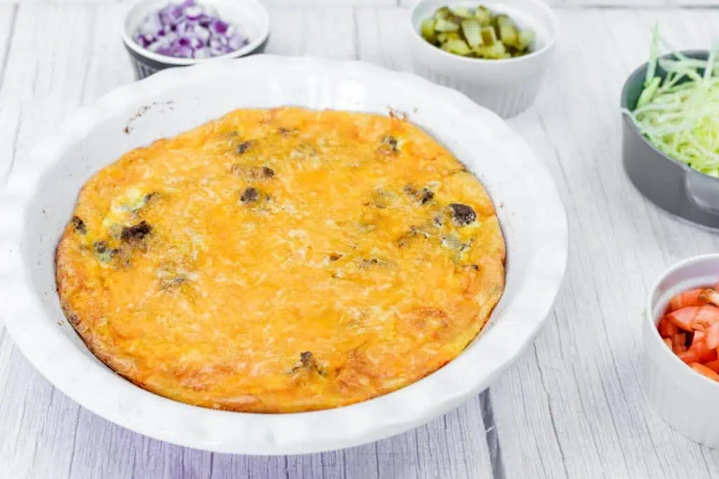 A baked cheeseburger pie with a golden-brown top in a white dish, surrounded by small bowls of chopped red onion, pickles, shredded lettuce, and diced tomato.