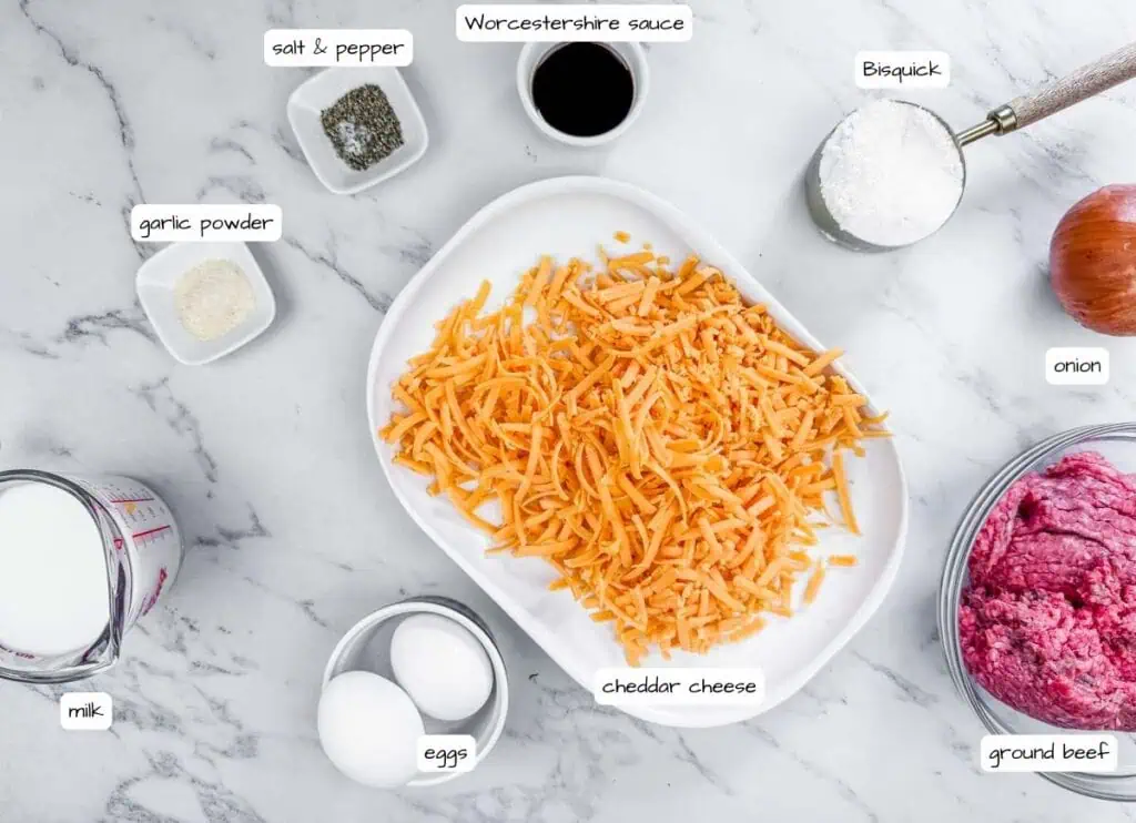 Ingredients for a recipe laid out on a marble counter: shredded cheddar cheese, ground beef, eggs, milk, Bisquick, onion, Worcestershire sauce, garlic powder, salt, and pepper.