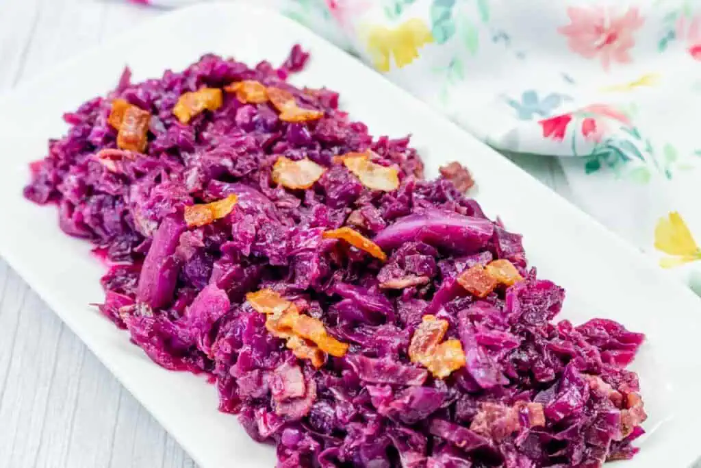 A rectangular white plate with cooked red cabbage mixed with pieces of bacon, placed on a light-colored table next to a floral-patterned cloth.