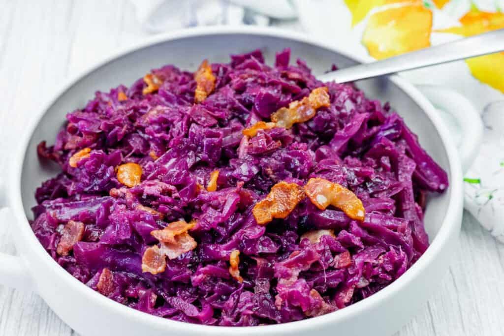 A bowl of cooked red cabbage mixed with pieces of bacon, served with a metal spoon.