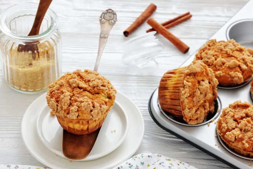 A cinnamon streusel muffin sits on a plate with a wooden spatula, next to a jar of sugar, cinnamon sticks, and a muffin tin holding more muffins.