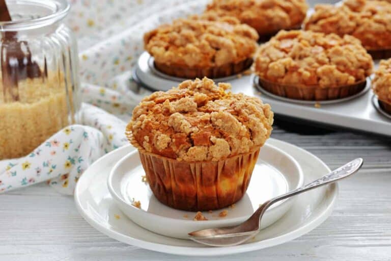 A crumb-topped muffin sits on a white plate with a spoon, with more muffins and a jar of brown sugar in the background.