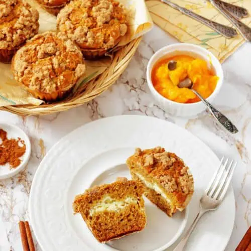 A plate with a halved streusel muffin, a fork, and a knife; more muffins in a basket and a bowl of orange spread are on the table, along with cinnamon sticks and forks.