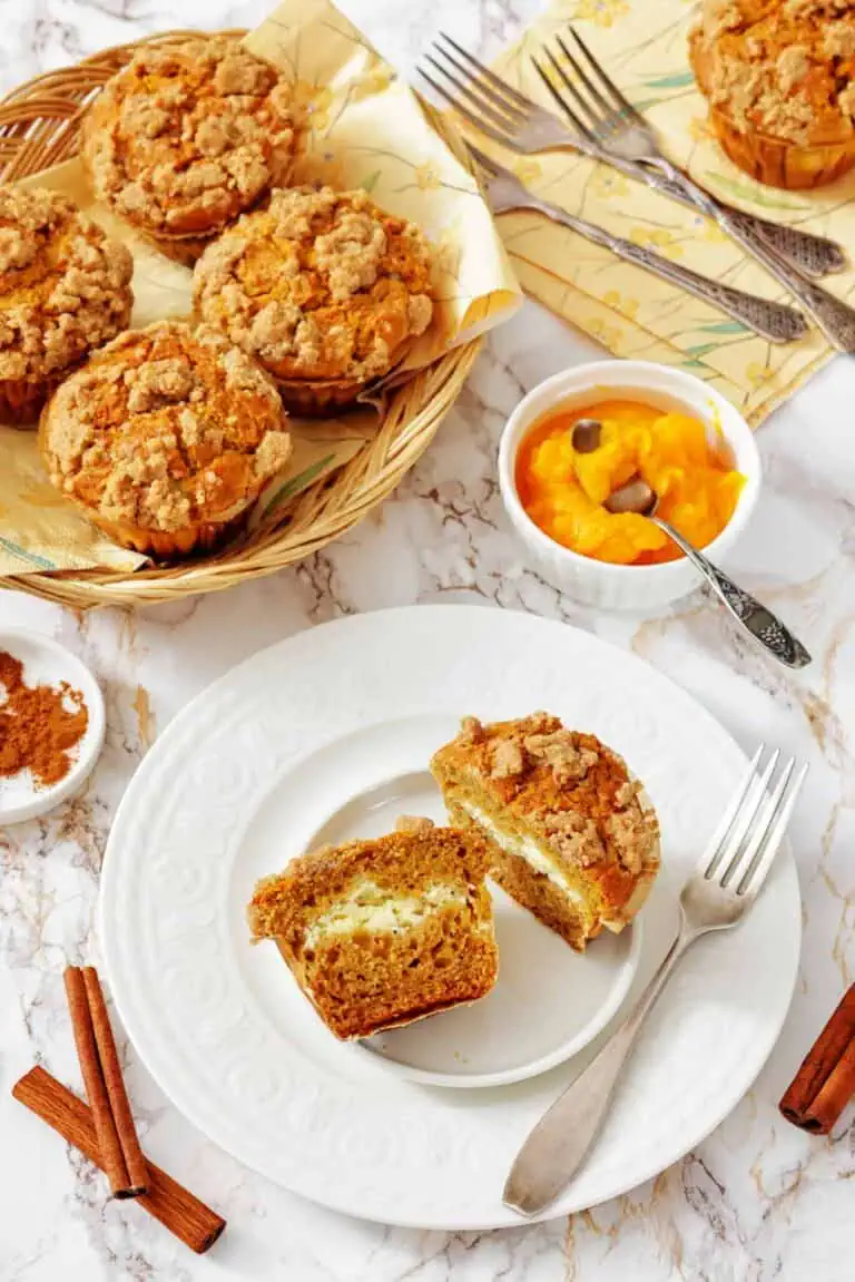 A plate with a halved streusel muffin, a fork, and a knife; more muffins in a basket and a bowl of orange spread are on the table, along with cinnamon sticks and forks.