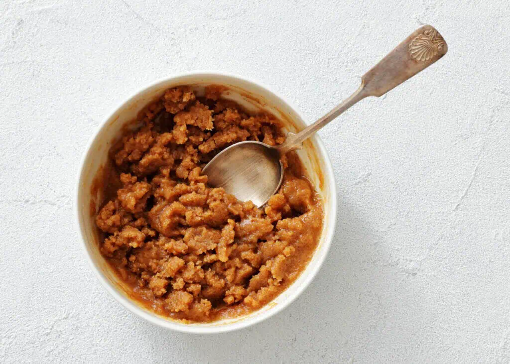 A bowl of brown, grainy paste with a silver spoon resting inside, placed on a white textured surface.