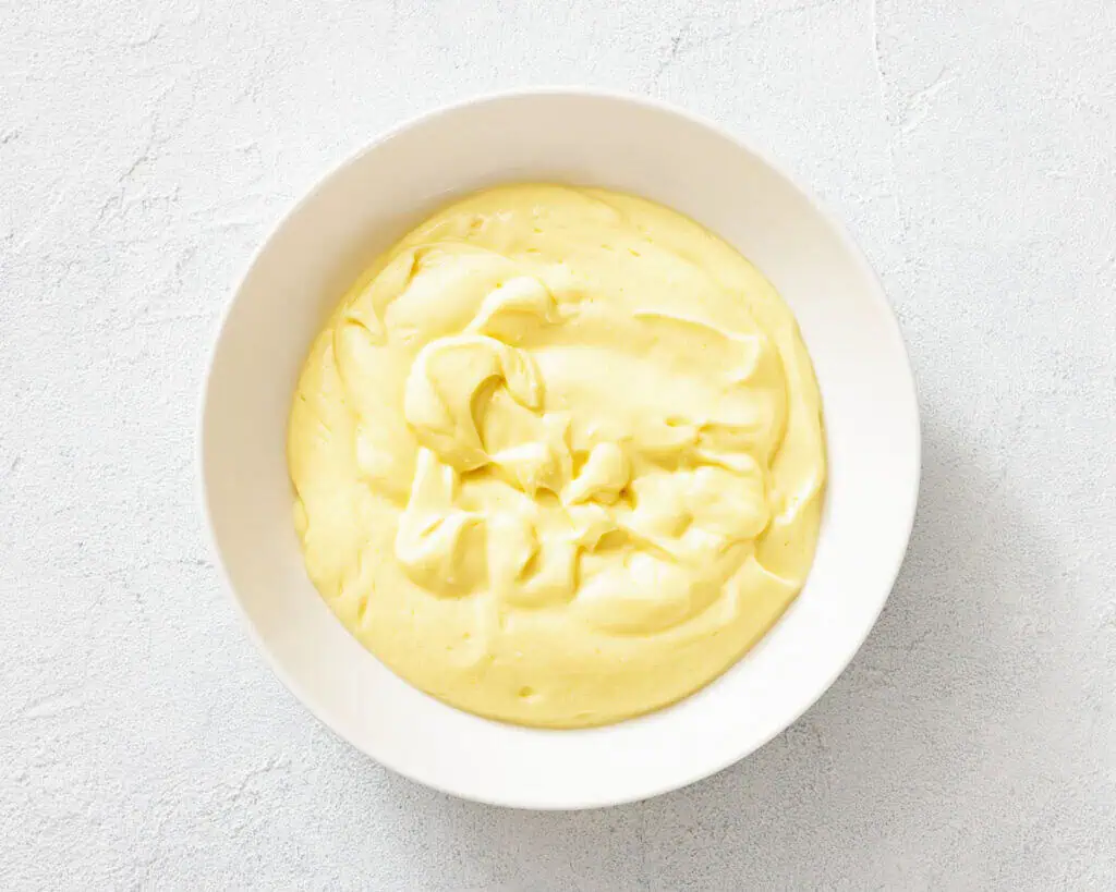 A white bowl filled with smooth, yellow mayonnaise on a light textured surface.